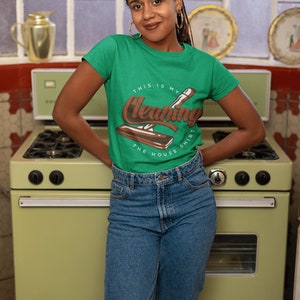 May include: A woman wearing a green t-shirt with the text "This is my cleaning shirt the house" and a brown illustration of a cleaning tool. She is standing in front of a vintage green stove.