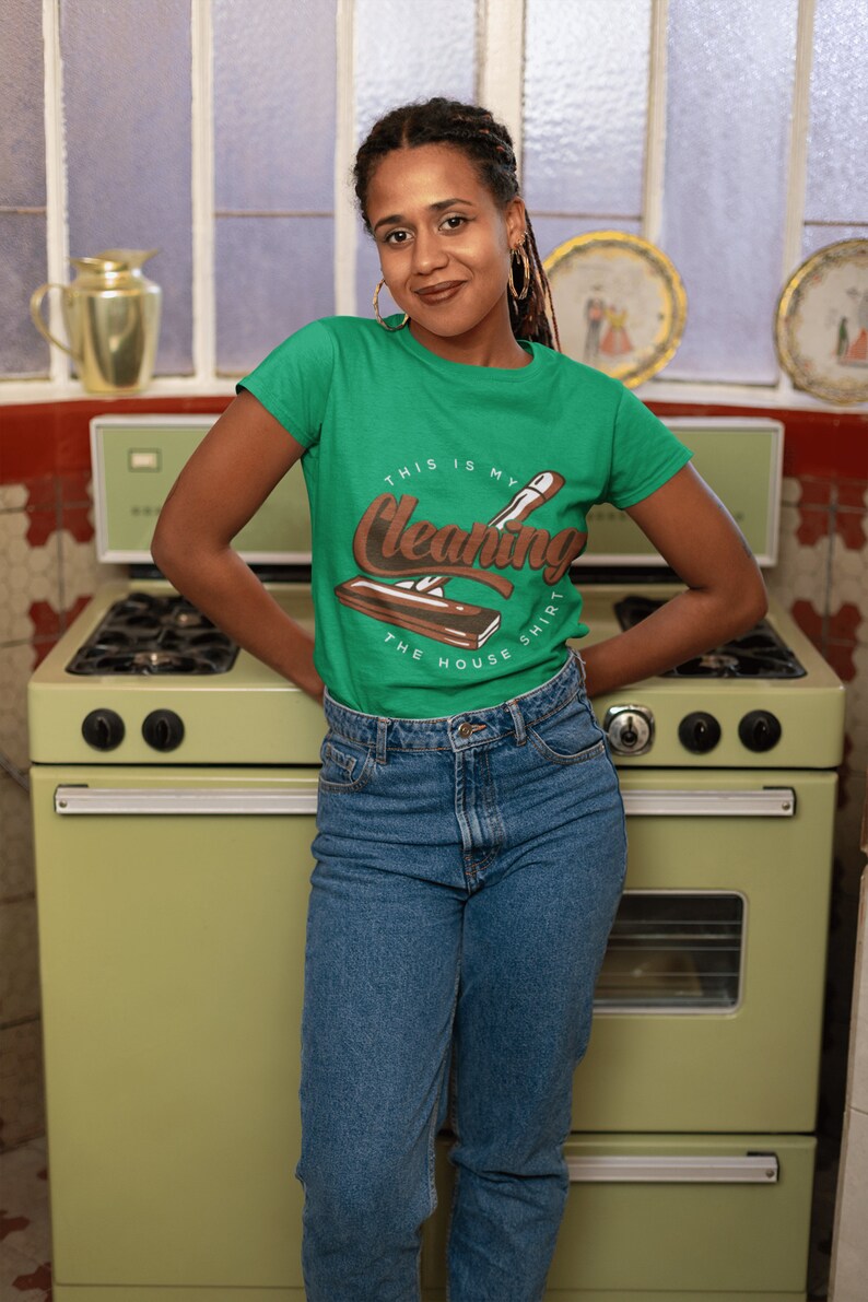 May include: A woman wearing a green t-shirt with the text "This is my cleaning shirt the house" and a brown illustration of a cleaning tool. She is standing in front of a vintage green stove.