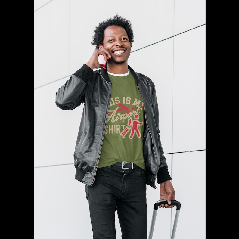 Puede incluir: Un hombre con una camiseta verde con el texto "This is my Airport Shirt" en una fuente de estilo vintage. Lleva una chaqueta de cuero negra y pantalones vaqueros negros. Est&aacute; hablando por tel&eacute;fono y sonriendo.