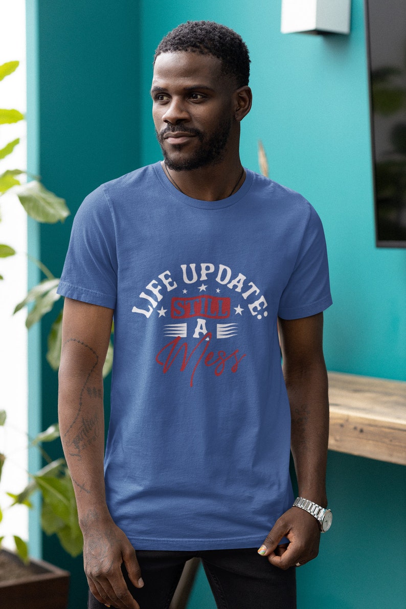 以下が含まれることがあります： 白い文字と赤い文字で「Life Update: Still a Mess」と書かれた青いTシャツ。テキストは星とストライプで囲まれています。