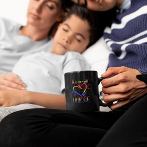 May include: A black coffee mug with a rainbow heart and the text "We are all UNIQUE" in white. The mug is being held by a person sitting on a bed with two other people sleeping behind them.
