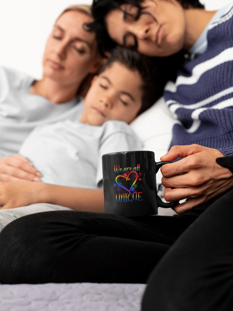 May include: A black coffee mug with a rainbow heart and the text "We are all UNIQUE" in white. The mug is being held by a person sitting on a bed with two other people sleeping behind them.
