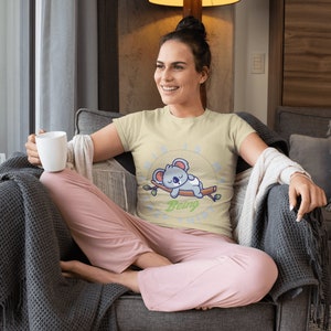 以下が含まれることがあります： 薄い黄色のTシャツを着た女性。Tシャツには、枝で眠るコアラの漫画と「Being Lazy」という文字がプリントされています。彼女は灰色のソファに座り、灰色の毛布をかけ、白いマグカップを持っています。