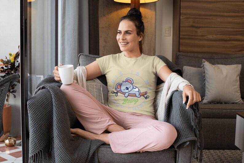 以下が含まれることがあります： 薄い黄色のTシャツを着た女性。Tシャツには、枝で眠るコアラの漫画と「Being Lazy」という文字がプリントされています。彼女は灰色のソファに座り、灰色の毛布をかけ、白いマグカップを持っています。