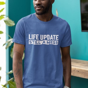 May include: A man wearing a blue t-shirt with the text "LIFE UPDATE STILL A MESS" printed in white.