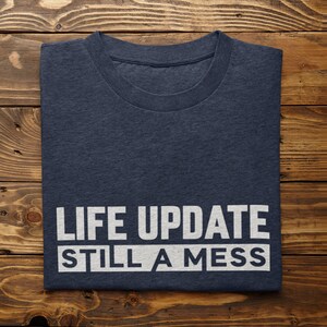 May include: A navy blue t-shirt with the text "LIFE UPDATE STILL A MESS" printed in white.