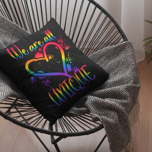 Puede incluir: Una almohada negra con un coraz&oacute;n arco&iacute;ris y el texto "We are all unique". La almohada est&aacute; sentada en una silla negra con una manta de color gris y blanco.