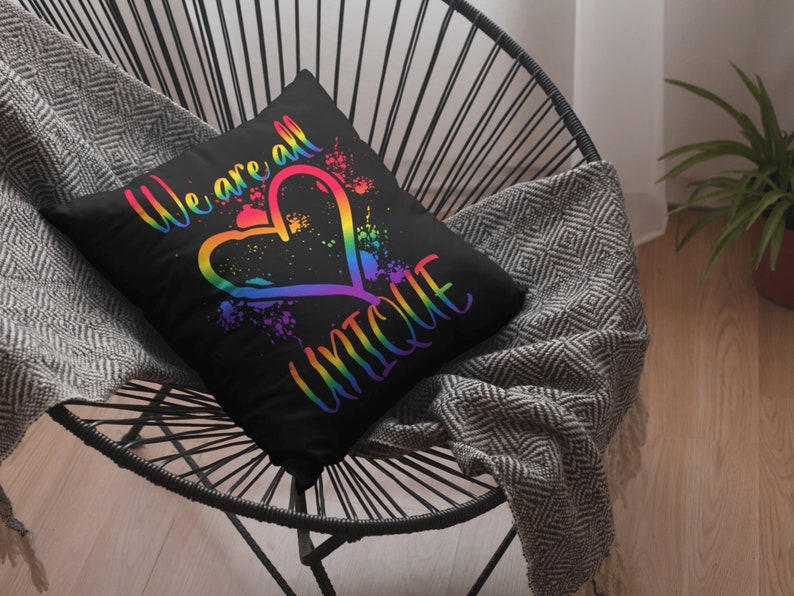 Puede incluir: Una almohada negra con un coraz&oacute;n arco&iacute;ris y el texto "We are all unique". La almohada est&aacute; sentada en una silla negra con una manta de color gris y blanco.