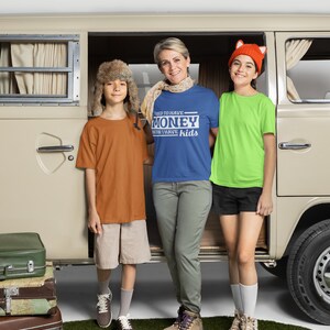 May include: A family of three standing in front of a vintage van. The woman is wearing a blue t-shirt with the text "I used to have money now I have kids". The boy on the left is wearing a brown t-shirt and a fur hat. The girl on the right is wearing a green t-shirt and a fox hat.