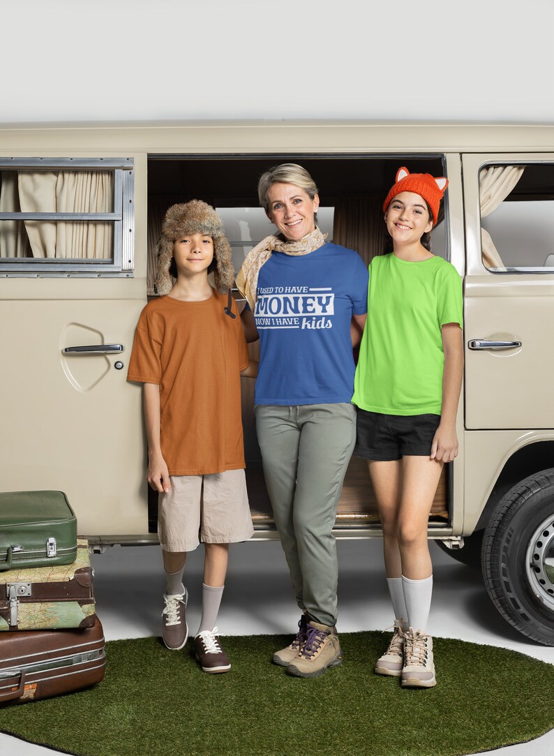 May include: A family of three standing in front of a vintage van. The woman is wearing a blue t-shirt with the text "I used to have money now I have kids". The boy on the left is wearing a brown t-shirt and a fur hat. The girl on the right is wearing a green t-shirt and a fox hat.
