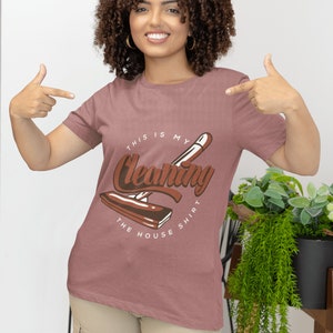 May include: A woman wearing a pink t-shirt with the text "This is my Cleaning Shirt" and a graphic of a cleaning tool. The shirt is being pointed to by the woman's hands.