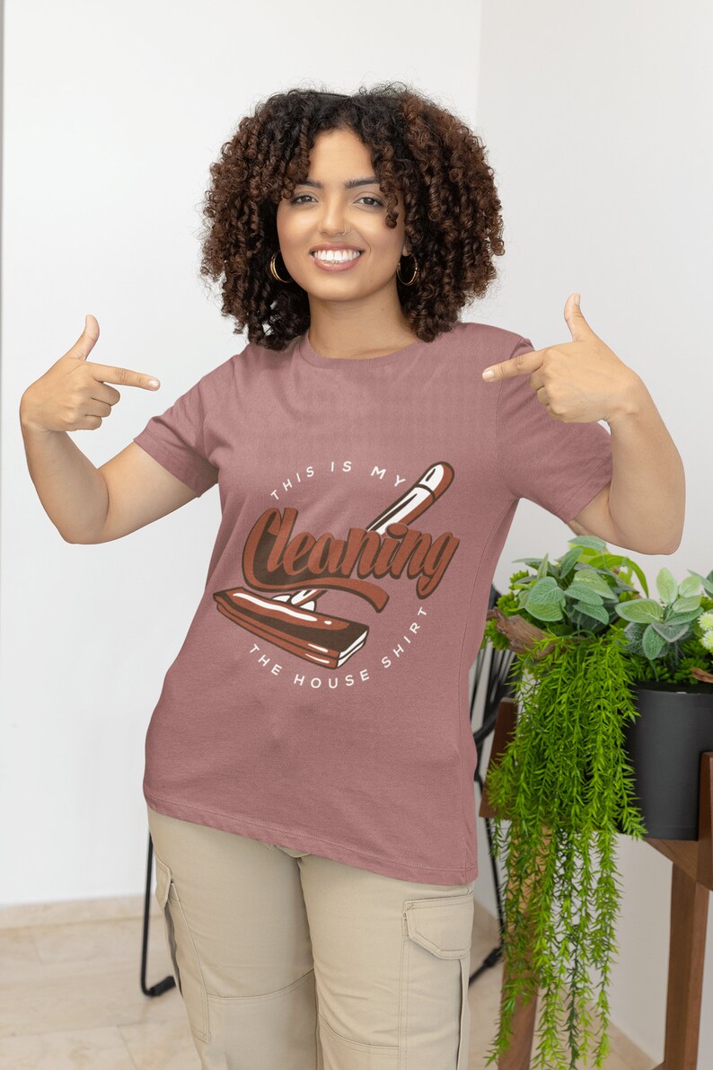 May include: A woman wearing a pink t-shirt with the text "This is my Cleaning Shirt" and a graphic of a cleaning tool. The shirt is being pointed to by the woman's hands.