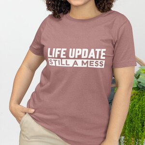 May include: A woman wearing a mauve t-shirt with the text "LIFE UPDATE STILL A MESS" printed in white. She is wearing sunglasses and has curly hair.