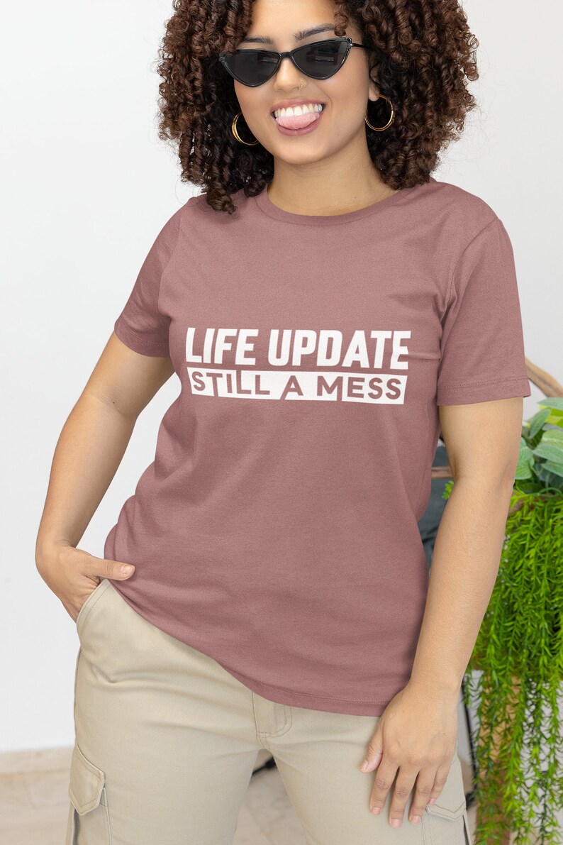 May include: A woman wearing a mauve t-shirt with the text "LIFE UPDATE STILL A MESS" printed in white. She is wearing sunglasses and has curly hair.