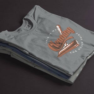 May include: A stack of gray t-shirts with a brown graphic that says "This is my cleaning the house shirt".