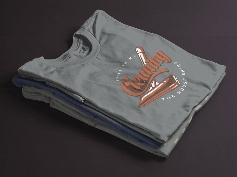 May include: A stack of gray t-shirts with a brown graphic that says "This is my cleaning the house shirt".