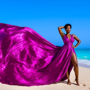 May include: A woman wearing a flowing, magenta-colored dress with a high slit stands on a sandy beach. The dress's fabric billows in the wind against a backdrop of a bright blue sky and turquoise water. The dress has a plunging neckline.
