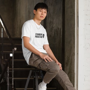 May include: A white t-shirt with a black logo that says "Strawberry Productions" and a small strawberry graphic. The person wearing the shirt is sitting on a stool.