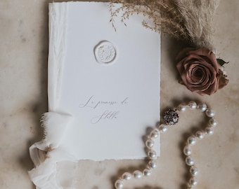 May include: A white paper booklet with deckled edges, tied with a frayed white ribbon, featuring a white wax seal and the text "Le promesse di Stella". Accompanied by a single mauve rose, dried pampas grass, and a strand of white pearls.