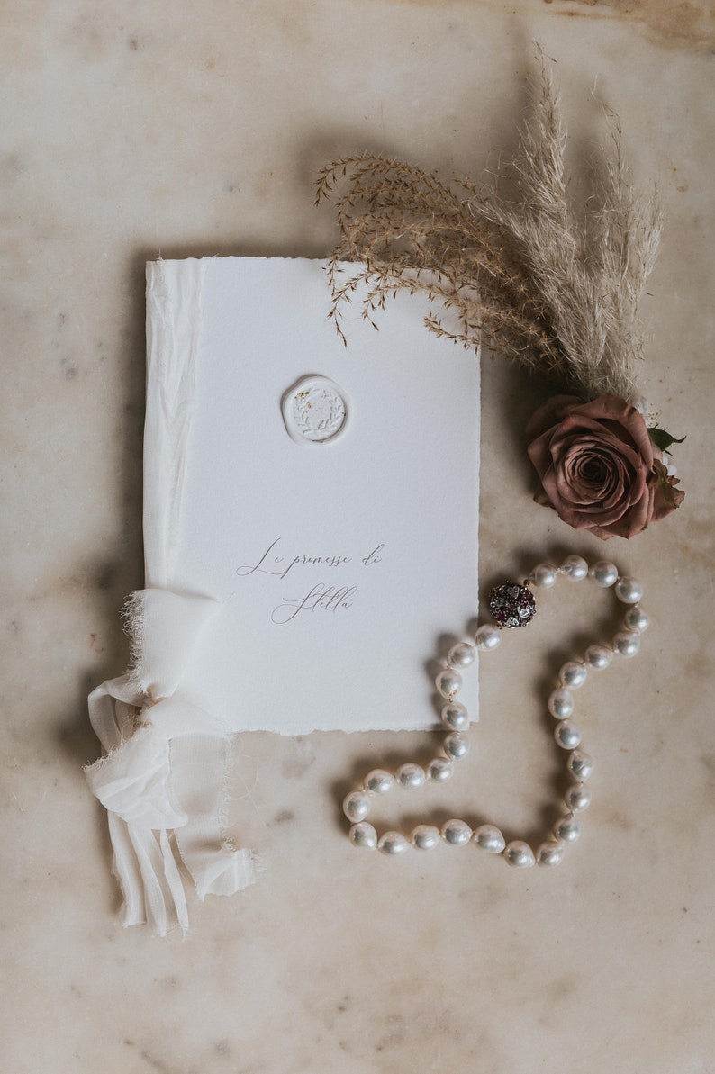 May include: A white card with the text "Le promesse de Stella" in cursive script. The card is decorated with a white wax seal and a white ribbon. A pearl necklace with a jeweled clasp and a single pink rose are also in the image.