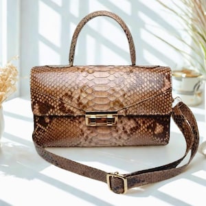 Könnte beinhalten: Eine braune Schlangenlederhandtasche mit einem goldenen Verschluss und einem abnehmbaren Schulterriemen. Die Tasche hat einen oberen Griff und einen Klappenverschluss.