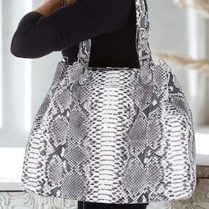 Op de afbeelding: Een grote, witte en grijze schlangenleren tote bag met zwarte banden en gespen.