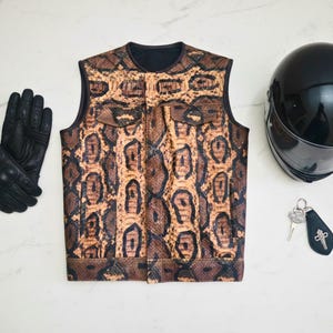 Könnte beinhalten: Eine Weste mit Schlangenmuster mit zwei Vordertaschen, schwarze Motorradhandschuhe, ein schwarzer Integralhelm und ein Schlüsselanhänger mit Kreuzanhänger. Die Artikel sind auf einer weißen Oberfläche angeordnet.