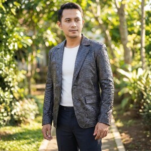 May include: A dark grey blazer with a textured pattern, worn over a white t-shirt and dark blue trousers. The blazer has a classic two-button design and a notched lapel. The person is standing outdoors in a garden setting.