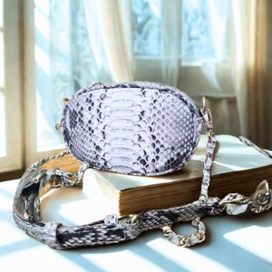 Könnte beinhalten: Eine weiße und graue Schlangenleder-Umhängetasche mit einer goldenen Kettenkette. Die Tasche liegt auf einem Buch, wobei der Riemen über die Seite gelegt ist.