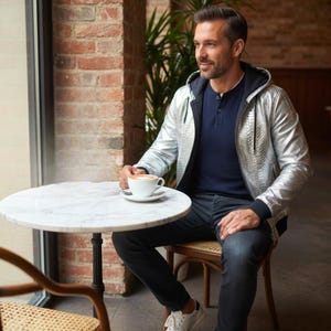 Puede incluir: Una persona con una chaqueta plateada metálica con capucha, camisa azul marino y vaqueros azul oscuro, sentada en una mesa con tapa de mármol. La persona sostiene una taza de café. El entorno parece ser un café o restaurante.