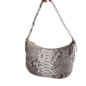 Op de afbeelding: Een klein, wit en grijs schlangenleren handtasje met een gebogen vorm en een enkele band.