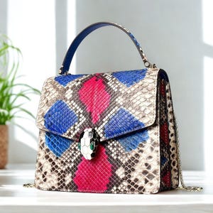 Pode incluir: Uma pequena bolsa de mão estruturada com alça superior e alça de corrente. A bolsa é feita de pele de cobra com um padrão azul, rosa e marrom. Um design de cabeça de cobra é apresentado na frente da bolsa.