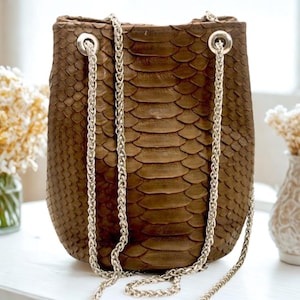 Puede incluir: Un bolso marrón tipo cubo con un patrón texturizado similar a la piel de serpiente. El bolso presenta una correa de cadena plateada y herrajes plateados. El bolso mide aproximadamente 25 cm de alto y 20 cm de ancho. El bolso está sobre una superficie blanca.