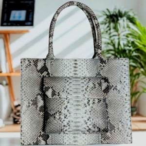 Puede incluir: Un bolso tote rectangular con un estampado de piel de serpiente en tonos grises y blancos. El bolso tiene dos asas redondeadas y un bolsillo delantero. El fondo incluye un escritorio, un portátil y plantas en macetas.