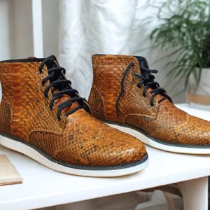 Könnte beinhalten: Ein Paar braune Lederstiefel mit Schlangenmuster. Die Stiefel haben schwarze Schnürsenkel, eine weiße Sohle und ein hohes Design. Sie werden auf einer weißen Oberfläche präsentiert.