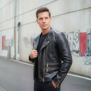 May include: A black leather jacket with a snakeskin pattern, featuring a classic asymmetrical zipper design and multiple zippered pockets. The jacket is worn over a black shirt. The background includes a concrete wall with graffiti.