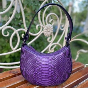 May include: A small, purple handbag made of snakeskin with a curved shape and a single strap.