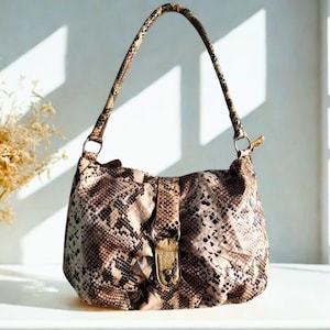 Puede incluir: Un bolso de hombro con estampado de piel de serpiente en tonos marrón y beige. Tiene un asa y un cierre dorado. El bolso está sobre una superficie blanca.