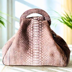 May include: A brown handbag with a rounded handle and a textured snakeskin pattern. The bag has a structured shape and a zip closure. The bag is on a white surface, with a window and plants in the background.