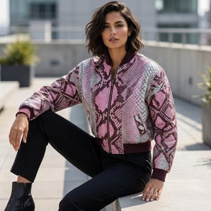 May include: A pink and gray snakeskin-patterned bomber jacket with a zipper closure. The jacket has a burgundy collar and cuffs. The person is wearing black pants and black boots.