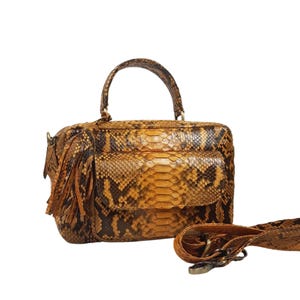 Puede incluir: Un bolso de mano de piel de serpiente marrón y amarillo con asa superior y correa de hombro desmontable. El bolso tiene un detalle de borla y un bolsillo frontal.