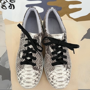 Könnte beinhalten: Ein Paar weiße Sneaker mit schwarzen Schnürsenkeln und einem Schlangenhautmuster.