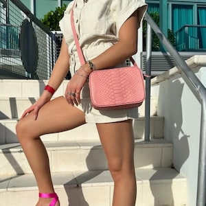 Op de afbeelding: Een roze slangleren schoudertas met een lange band. De tas wordt gedragen door iemand die op een trap staat.