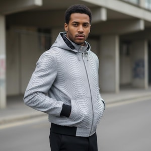 Pode incluir: Casaco cinza claro com capuz e padrão de pele de cobra. O casaco tem fecho de correr, punhos pretos e cós preto. A pessoa está na rua com um edifício ao fundo.