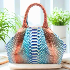 Puede incluir: Un bolso colorido con un patrón único de piel de serpiente. El bolso presenta una combinación de colores azul, naranja y marrón con una base blanca y un asa marrón. El bolso está sobre una superficie blanca.