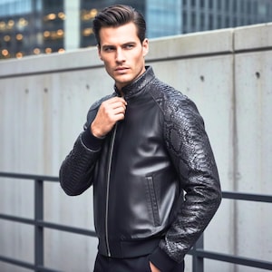 May include: Black leather jacket with snake-skin textured sleeves and shoulder accents. The jacket has a zip-up front and a bomber-style collar. The model is wearing a black jacket and black trousers.