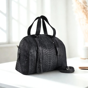 Pode incluir: Bolsa de mão de couro preto com textura de pele de cobra, com alça superior e alça de ombro removível. A bolsa tem formato arredondado, detalhe de borla e fecho de zíper. A bolsa está em uma superfície branca.