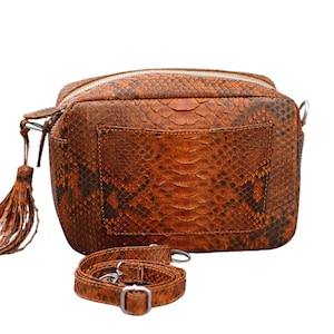 Könnte beinhalten: Eine braune Schlangenleder-Umhängetasche mit Reißverschluss und Quaste. Die Tasche hat einen langen, verstellbaren Riemen und ein kleines Fach auf der Rückseite.