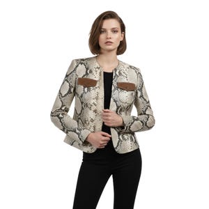 Puede incluir: Chaqueta con estampado de piel de serpiente con detalles marrones. La chaqueta tiene un diseño sin cuello y dos pequeños bolsillos con detalles de cuero marrón. La modelo lleva un top negro y pantalones negros. La chaqueta es de color neutro con estampado de reptil.