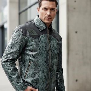 Op de afbeelding: Een groene en zwarte leren jas met een slangenpatroon. De jas heeft een ritssluiting en een kraag. De schouders en kraag zijn zwart, terwijl de body van de jas groen is. Het model staat voor een betonnen gebouw.